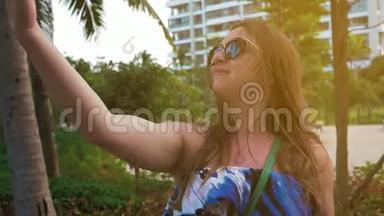 漂亮的快乐女孩戴着眼镜和蓝色连衣裙，在手机上摆出自拍。 绿色的手掌和美丽的阳光。 中等